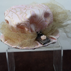 May include: A pink and gold hat with a small, decorative book charm. The hat is made of a patterned fabric and has a gold tulle bow.
