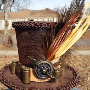 May include: A brown leather top hat with a steampunk design. The hat has a gear and a metal arrow on the front, and a brown and yellow feather on the top.