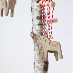 May include: Three brown wooden horse ornaments hanging from a birch tree branch with red and white twine.
