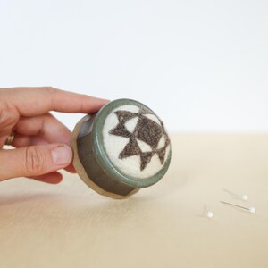 May include: A round pincushion featuring a dark green ceramic base and a white felted top with a dark brown geometric star pattern. The pincushion has a smooth, glazed finish on the sides and a soft, textured surface on the top for holding pins.