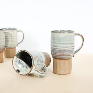 May include: Several handmade ceramic mugs with a light blue and brown striped design. The mugs have a tan base and a curved handle. One mug is lying on its side, revealing the interior.
