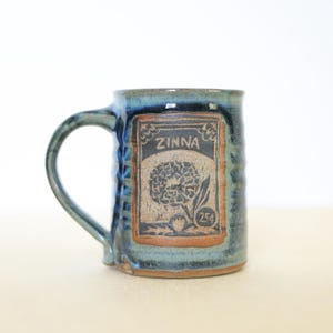 May include: A ceramic mug with a blue and green glazed finish featuring a rectangular stamp on the front. The stamp depicts a flower with the text ZINNA and 25 cents. The mug has a curved handle and a textured, slightly rippled cylindrical body.