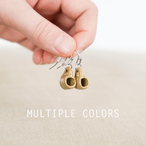 May include: A pair of small, tan-colored ceramic earrings with a unique, rounded shape, held up by silver-colored hooks. The earrings have a hollowed-out design. The text "MULTIPLE COLORS" is displayed at the bottom of the image.