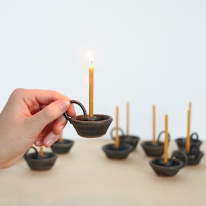 May include: A small, dark brown ceramic candle holder with a handle, holding a lit yellow candle. Several similar candle holders are arranged in the background. The candle flame is bright against the neutral background.