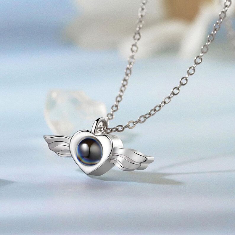 May include: A silver heart-shaped pendant necklace with small wings on either side. The heart has a dark, round center. The necklace chain is also silver and has a delicate link design. The background is a soft, light blue.