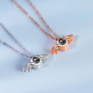 May include: Two necklaces with heart-shaped pendants and wing accents. One necklace is silver, the other is rose gold. Each pendant has a dark, round center. The chains are delicate and the background is a light blue.
