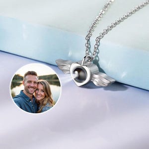 May include: A silver-colored heart-shaped pendant necklace with attached wings. The pendant has a small, round opening in the center. A photo of a couple is in the lower left corner. The necklace hangs on a silver chain.