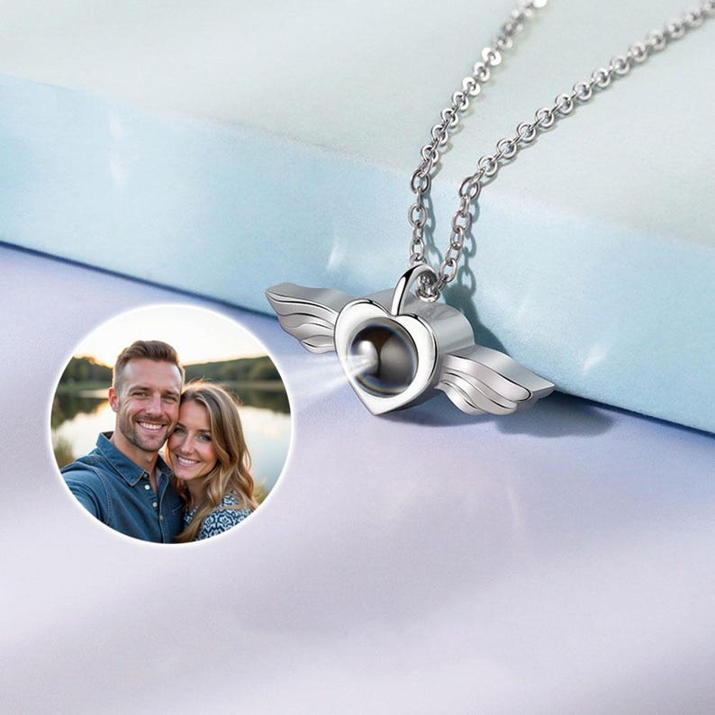 May include: A silver-colored heart-shaped pendant necklace with attached wings. The pendant has a small, round opening in the center. A photo of a couple is in the lower left corner. The necklace hangs on a silver chain.