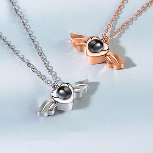 May include: Two necklaces with heart-shaped pendants and wing accents. One necklace is silver, the other is rose gold. Each pendant has a dark, spherical center. The chains are delicate and the background is a light blue.