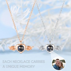 May include: Two necklaces with heart-shaped pendants and wing accents. One is rose gold, the other silver. Each pendant has a dark center. The text reads: "EACH NECKLACE CARRIES A UNIQUE MEMORY."