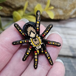 Peut inclure: Une épingle en émail noire, jaune et rouge représentant une araignée avec un design détaillé. L'araignée a huit pattes et un corps rond avec un motif jaune et brun.