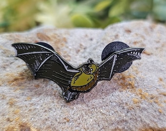 Puede incluir: Un pin de esmalte negro y dorado de un murciélago en vuelo. El murciélago tiene un diseño detallado con alas negras y un cuerpo dorado.