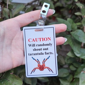 Pode incluir: Uma placa retangular branca com um clipe de plástico transparente. A placa diz "CAUTION" em vermelho, seguido de texto sobre gritar fatos sobre tarântulas, e apresenta uma ilustração de tarântula laranja e azul.
