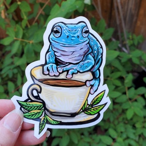 May include: A blue frog with a white belly sits on the rim of a white teacup with a brown liquid inside. Green leaves surround the teacup and frog.