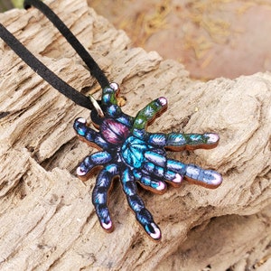 May include: A wooden pendant necklace featuring a colorful tarantula design. The spider has a vibrant mix of blue, purple, pink, and green hues. The pendant is suspended from a black cord necklace, creating a unique and eye-catching accessory.