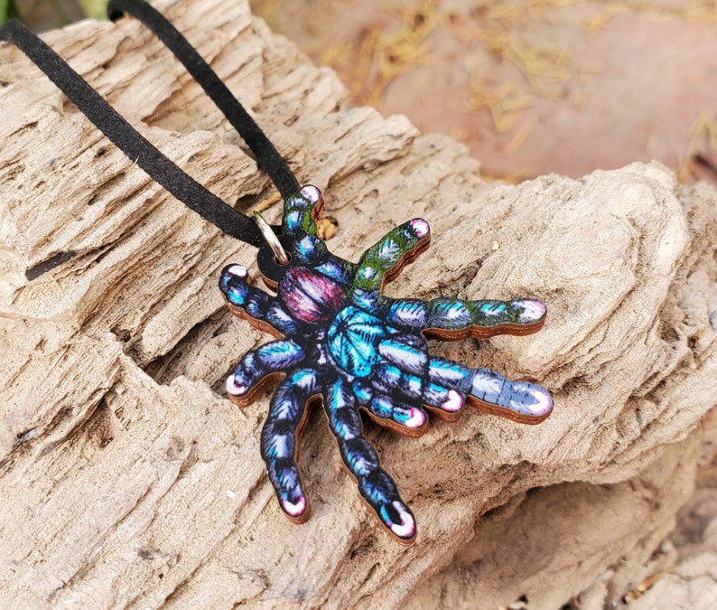 May include: A wooden pendant necklace featuring a colorful tarantula design. The spider has a vibrant mix of blue, purple, pink, and green hues. The pendant is suspended from a black cord necklace, creating a unique and eye-catching accessory.