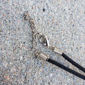 May include: Close-up of a black suede necklace with a silver-colored chain and clasp. The necklace is lying on a gray, textured surface. The chain has a heart-shaped design.