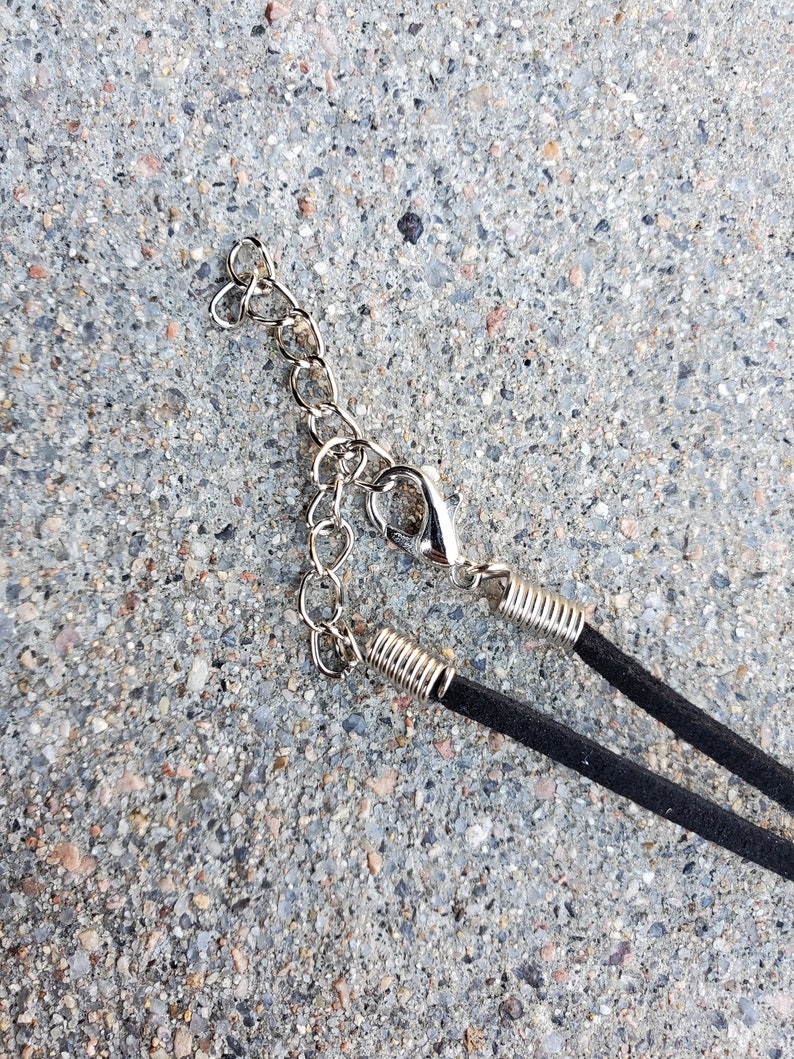 May include: Close-up of a black suede necklace with a silver-colored chain and clasp. The necklace is lying on a gray, textured surface. The chain has a heart-shaped design.