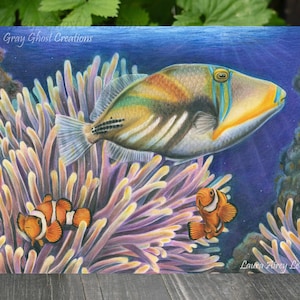 May include: A colorful illustration of a fish swimming near coral and two clownfish. The fish has yellow, orange, and blue markings. The coral is purple and white. The background is blue. The artwork is signed "Laura Airey Le".