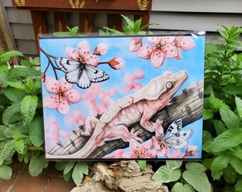 Könnte beinhalten: Ein Aquarellbild eines rosa-weißen Crested Geckos auf einem Ast mit rosa Kirschblüten und weißen Schmetterlingen. Der Gecko sitzt auf einem Ast mit seinem Schwanz um den Ast gewickelt. Die Kirschblüten stehen in voller Blüte und die Schmetterlinge fliegen um den Gecko herum.