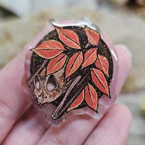 May include: A clear resin pin featuring a brown bat with pink ears and wings, surrounded by red and orange leaves with gold glitter accents.