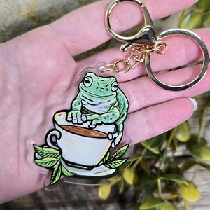 Puede incluir: Un llavero de rana verde con una taza de té. La rana está sentada en el borde de la taza y la sostiene con sus manos. El llavero está hecho de acrílico transparente y tiene una cadena dorada.