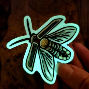 May include: A glow-in-the-dark sticker of a moth with detailed wings and a segmented body. The moth is depicted in shades of green, blue, and white.