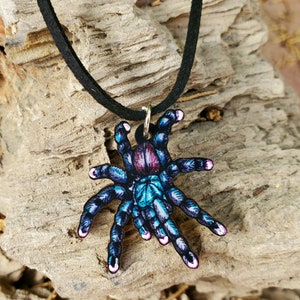May include: A pendant necklace featuring a colorful tarantula design. The spider is depicted in shades of blue, teal, and purple, with a black cord. The pendant is attached to a black cord necklace.