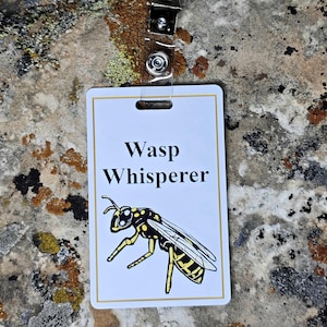 May include: A white rectangular badge with the words "Wasp Whisperer" in black text, and a detailed illustration of a wasp in yellow and black. The badge is attached to a clear plastic holder with a metal clip.