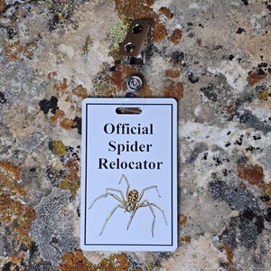 Może przedstawiać: Biała odznaka z napisem "Official Spider Relocator" i grafiką pająka. Posiada metalowy klips i przezroczysty plastikowy pasek. Tło stanowi teksturowana powierzchnia skalna.