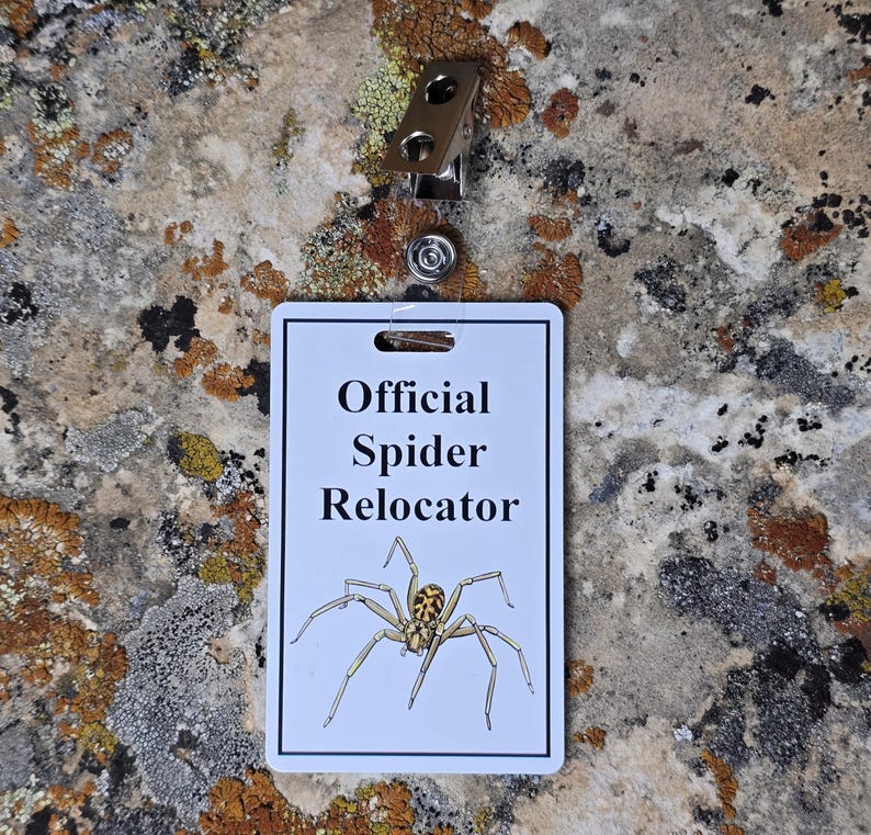 以下が含まれることがあります： 「Official Spider Relocator」の文字とクモのグラフィックが入った白いバッジ。金属製のクリップと透明なプラスチックストラップが付いています。背景は質感のある岩の表面です。