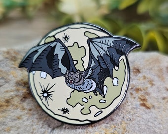 Puede incluir: Un pin de esmalte negro y gris de un murciélago volando frente a una luna amarilla con marcas grises y blancas.