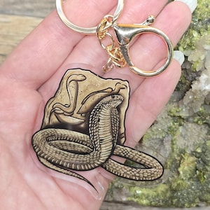May include: A gold-colored keychain with a clear acrylic charm featuring a detailed illustration of a cobra snake. The snake is depicted in shades of brown and tan, coiled in front of a rock formation. The keychain has a lobster clasp and a split ring.