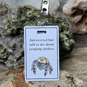 May include: A white rectangular badge with the text "Introverted but talk to me about jumping spiders." features a cartoon jumping spider illustration. The badge is attached to a metal clip with a clear plastic strap.