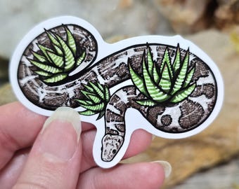 May include: A sticker featuring a brown and black snake with green succulent plants growing on top of it. The snake is curled up in a circle and the succulents are in full bloom.