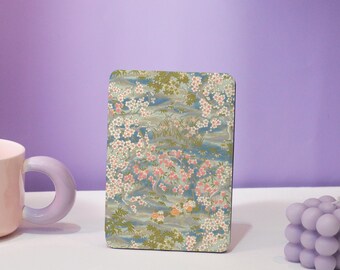 May include: Rectangular decorative item with a floral pattern. The design features pink and white blossoms, green foliage, and a blue and gray background. The item is standing upright on a white surface against a purple backdrop.