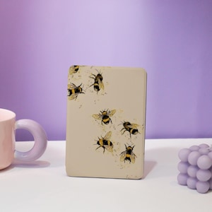 May include: A rectangular, off-white case featuring watercolor-style illustrations of bees in flight. The bees are black and yellow with delicate wing details. The case is set against a soft purple background, with a pink mug and a lavender candle.