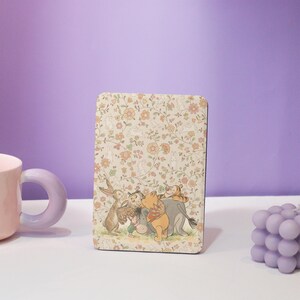 May include: A rectangular decorative item with a floral pattern and Winnie the Pooh characters. The design includes a rabbit, owl, Pooh Bear, Tigger, and Eeyore. The background is cream with orange and yellow flowers.