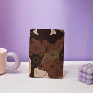 May include: A brown and beige camouflage pattern card holder with the Louis Vuitton logo.