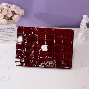May include: A red laptop skin with a crocodile pattern. The skin is glossy and has a white Apple logo in the center.
