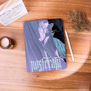 May include: A notebook with a fantasy illustration featuring two figures in shades of purple and blue, with the word "NOSTERATU" in a stylized font. A white stylus and a small candle are also visible.