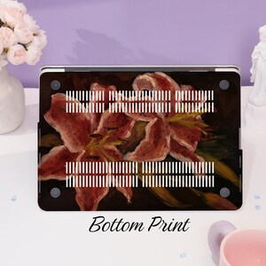 Peut inclure: Une coque d'ordinateur portable avec un imprim&eacute; floral repr&eacute;sentant des lys roses sur un fond sombre. La coque porte l'&eacute;tiquette "Bottom Print".