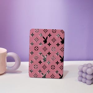 May include: Pink rectangular case featuring a repeating pattern of black bunny silhouettes, four-leaf clovers, and diamond shapes. Some shapes have silver glitter accents. The case is on a white surface with a purple background.
