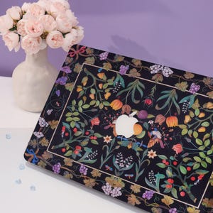 May include: A black laptop case with a floral design featuring a variety of flowers, leaves, and grapes. The design is in shades of pink, purple, green, and yellow. The Apple logo is visible on the case.