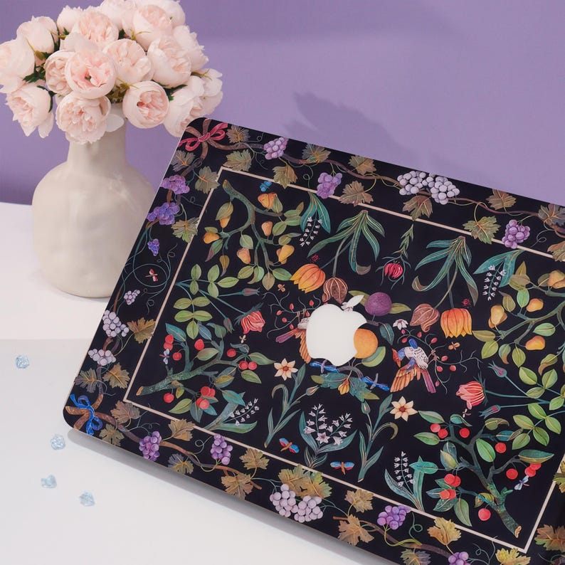 May include: A black laptop case with a floral design featuring a variety of flowers, leaves, and grapes. The design is in shades of pink, purple, green, and yellow. The Apple logo is visible on the case.