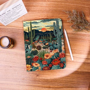 Pode incluir: Um caderno com uma ilustração de paisagem desértica com cactos, flores e um pôr do sol. A capa tem um design colorido com tons de verde, laranja e azul. Uma caneta stylus branca e uma pequena vela estão ao lado do caderno.