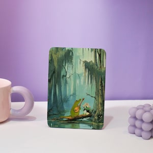 Op de afbeelding: Rechthoekige kunstdruk met een moeraslandschap met een groene alligator die banjo speelt en een kleine vogel met een hoed. Het kunstwerk speelt zich af in een moeras met hangend mos en een lichtgroen en blauw kleurenpalet.