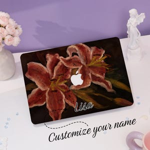 Peut inclure: Une peau d'ordinateur portable avec une peinture de deux lys rouges sur un fond sombre. La peau a un logo Apple blanc et le nom "Lisa" est imprim&eacute; en bas &agrave; droite. Le texte "Personnalisez votre nom" est imprim&eacute; sous la peau de l'ordinateur portable.