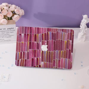 May include: A laptop case with a vibrant abstract design in shades of pink, purple, and yellow. The case features a white Apple logo and a textured, brushstroke-like pattern. The case is a protective accessory for a laptop.