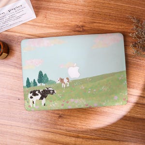 May include: A laptop hard shell case featuring a light green landscape illustration with two cows, green trees, and pink clouds. The design includes a central cutout for the Apple logo, showcasing a pastoral scene with a black and white cow.